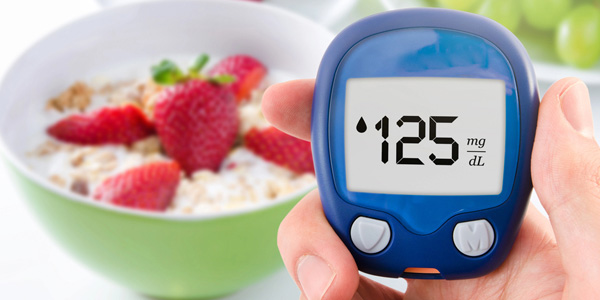 Hand hält ein blaues Blutzuckermessgerät mit Anzeige 125 mg/dL. Im Hintergrund steht eine grüne Schüssel mit Müsli und Erdbeeren, unscharf.